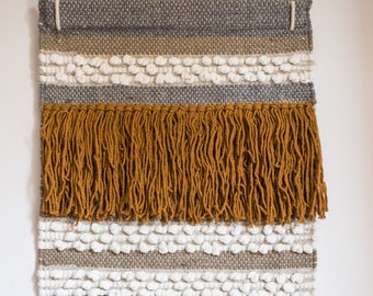 May include: A rectangular woven wall hanging featuring horizontal bands of gray, tan, and white yarn. The piece includes sections of textured white bobble knots, a thick layer of mustard yellow fringe, and a long white fringe at the bottom edge.
