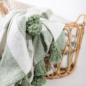 May include: A white and green woven throw blanket with tassels. The blanket is folded and draped over a wicker basket.