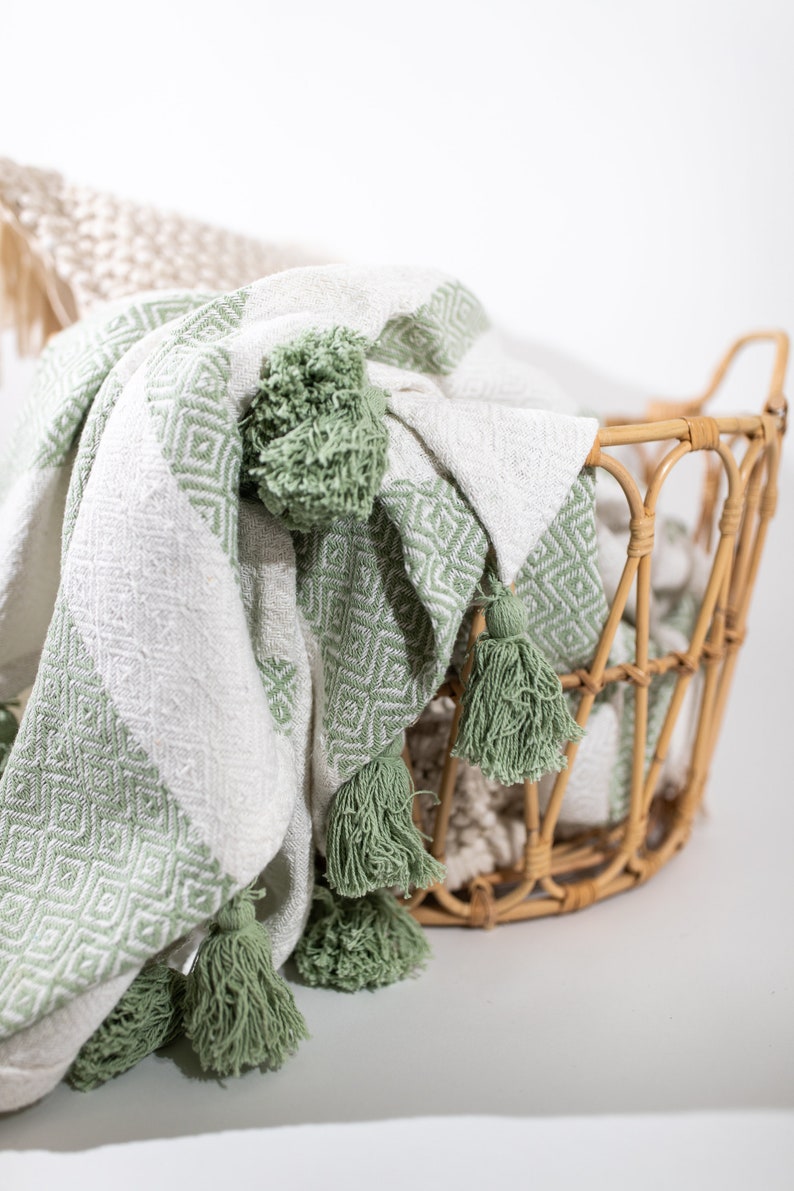 May include: A white and green woven throw blanket with tassels. The blanket is folded and draped over a wicker basket.