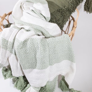 May include: A white and sage green throw blanket with a diamond pattern and tassels. The blanket is folded and draped over a wicker chair.