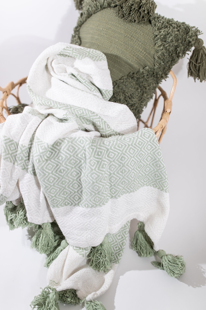 May include: A white and sage green throw blanket with a diamond pattern and tassels. The blanket is folded and draped over a wicker chair.