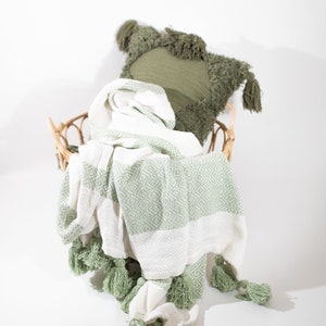 May include: A white and green throw blanket with tassels, folded and placed in a wicker basket with a green throw pillow.