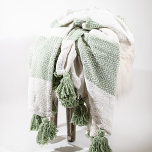 May include: A white and green throw blanket with tassels. The blanket is folded and draped over a stool.