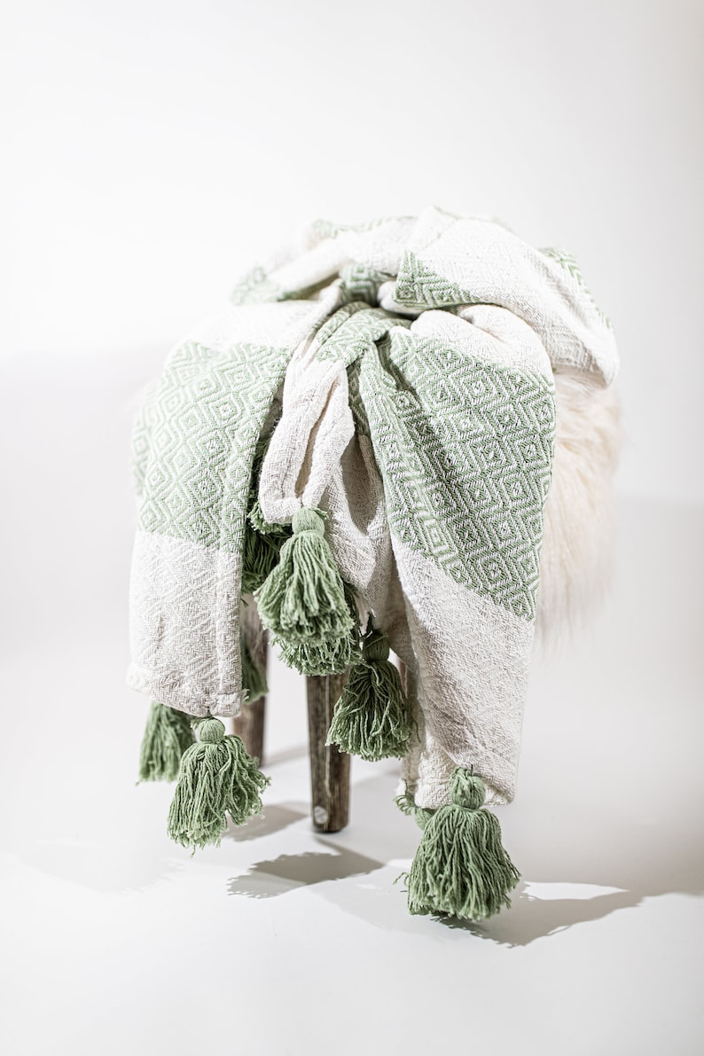 May include: A white and green throw blanket with tassels. The blanket is folded and draped over a stool.
