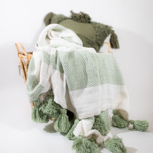 May include: A white and green throw blanket with tassels. The blanket is folded and placed in a wicker basket.