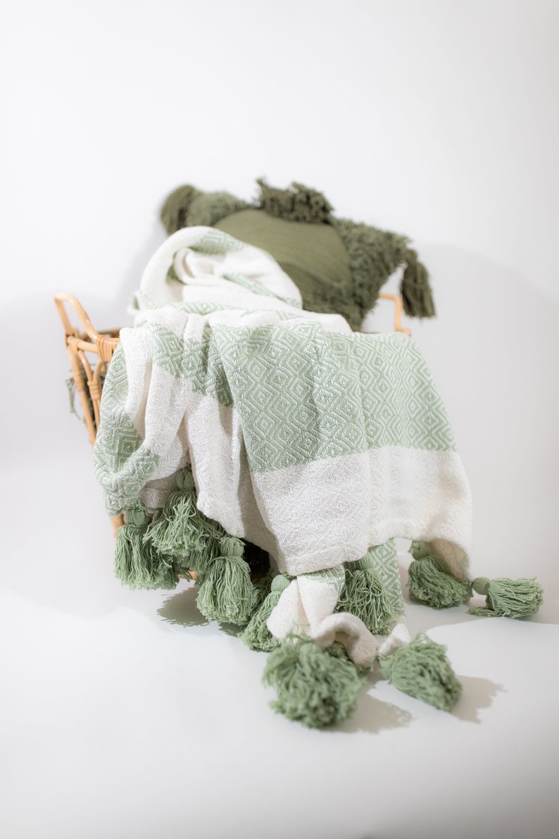 May include: A white and green throw blanket with tassels. The blanket is folded and placed in a wicker basket.