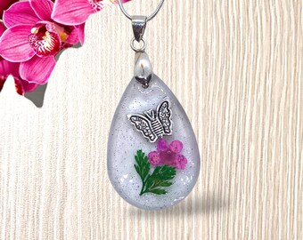 Pendentif femme ovale goutte d'eau, incrustation d'une fleur séchée et d'un papillon argenté dans un écrin de résine transparente pailletée