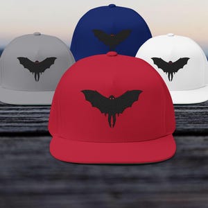 May include: A collection of flat-brimmed baseball caps in various colors: red, navy blue, white, and gray. Each cap features a black bat graphic on the front. The caps are displayed on a dark wooden surface.