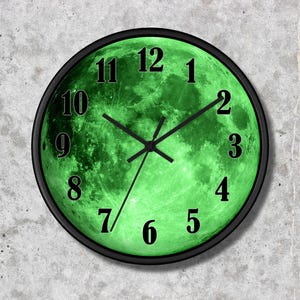 May include: A round wall clock with a black frame and a bright green lunar surface graphic on the face. The clock features black Arabic numerals from one to twelve and three black hands indicating the time against the detailed moon texture.