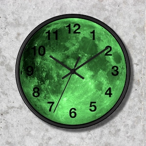 Puede incluir: Un reloj de pared redondo con un marco negro y una esfera verde con forma de luna. Las manecillas del reloj son negras y los números están en negrita negra. El reloj está sobre una pared gris texturizada.