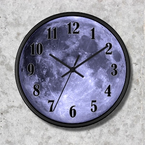May include: A round wall clock featuring a high-resolution image of the full moon as the clock face. The moon displays shades of blue and grey with visible craters. It has a thin black frame, black numbers from 1 to 12, and three black hands.
