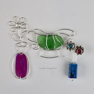 Peut inclure: Pièces d'art en fil de fer faites à la main représentant une fleur, une saucière avec une pierre verte et deux breloques en forme de fleur. La fleur a une pierre violette dans un vase. Les breloques ont des pierres bleues et rouges. Le nom de l'artiste, Heather Boyd Wire, est visible.