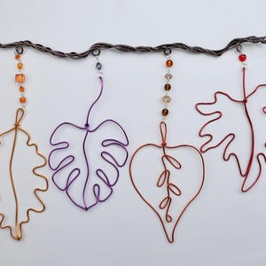 May include: A wall hanging featuring a dark twisted wire branch from which four distinct wire leaf shapes are suspended. The leaves are colored orange, purple, copper, and red, each accented with vertical strands of small colorful beads.