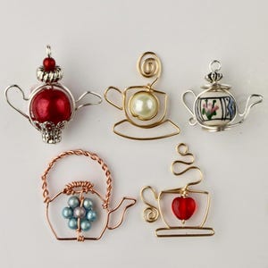 May include: Five handcrafted wire tea-themed ornaments are displayed. The collection includes teacups and teapots, some with red, white, and blue beads, and others with pearl accents. The ornaments are made of silver, gold, and copper-coloured wire.
