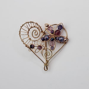 May include: A handcrafted gold-toned wire heart-shaped brooch. The brooch features a floral design with purple beads and accents. The heart shape has a spiral design on the left side and a loop at the bottom.
