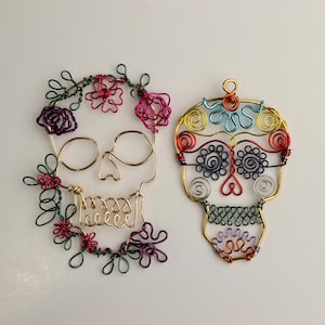 May include: Two wire-wrapped sugar skull ornaments. The skull on the left has a floral design with pink, purple, and green wire. The skull on the right has a colorful design with red, orange, yellow, blue, green, and purple wire.