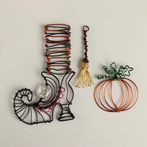 Pode incluir: Três ornamentos de Halloween envoltos em arame: uma bota de bruxa com um design vermelho e preto, uma vassoura dourada e uma abóbora com um caule verde.