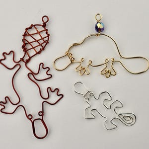 May include: Four wire animal ornaments: a red platypus, a gold platypus with a purple bead, a red lizard, and a silver lizard. The ornaments are handmade and have a whimsical, artistic design. The platypus ornaments have a loop for hanging.