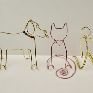Könnte beinhalten: Drei Drahtskulpturen von Tieren: ein goldener Hund mit einem Regenbogen-Perlenhalsband, eine rosafarbene Katze mit einem Herzen und eine goldene Strichmännchenfigur.