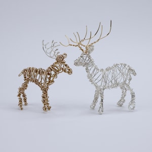 May include: Two wire sculptures of deer standing side by side. One deer is crafted from gold-toned wire, while the other is made from silver-toned wire. Both figures feature intricate, open-work wire bodies and detailed antlers extending upwards.