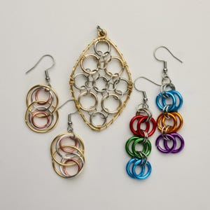 Könnte beinhalten: Ein Set aus drei Schmuckstücken aus Metalldraht. Der Anhänger ist oval geformt und hat ein Muster aus ineinandergreifenden Kreisen. Die Ohrringe bestehen beide aus mehreren Kreisen, ein Paar ist silber und gold, das andere ist regenbogenfarben.
