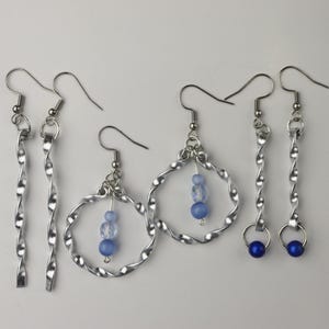 May include: Three pairs of silver earrings with blue beads. One pair features a twisted silver hoop with three blue beads. Another pair has a twisted silver dangle with a blue bead at the end. The third pair has a twisted silver dangle.
