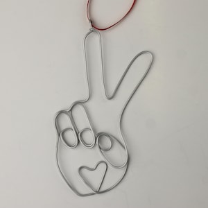 May include: A silver wire peace sign ornament with a small heart in the center. The ornament is attached to a red ribbon.