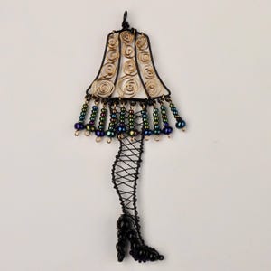 May include: A wire sculpture of a lampshade with a leg and a high heel shoe. The lampshade is made of gold wire and has a fringe of iridescent beads. The leg is made of black wire and is wrapped in a black net pattern. The shoe is black with iridescent beads.