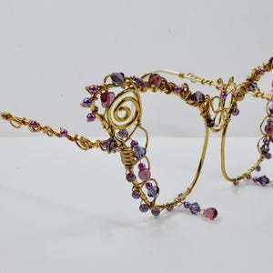 May include: Decorative glasses frames constructed from gold-toned wire, featuring intricate spiral patterns at the temples and embellished with small purple and lavender beads and faceted crystals along the rims and arms of the eyewear.