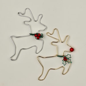 Peut inclure: Deux décorations de rennes en fil de fer, l'une argentée et l'autre dorée, avec des accents de baies rouges et de houx vert.