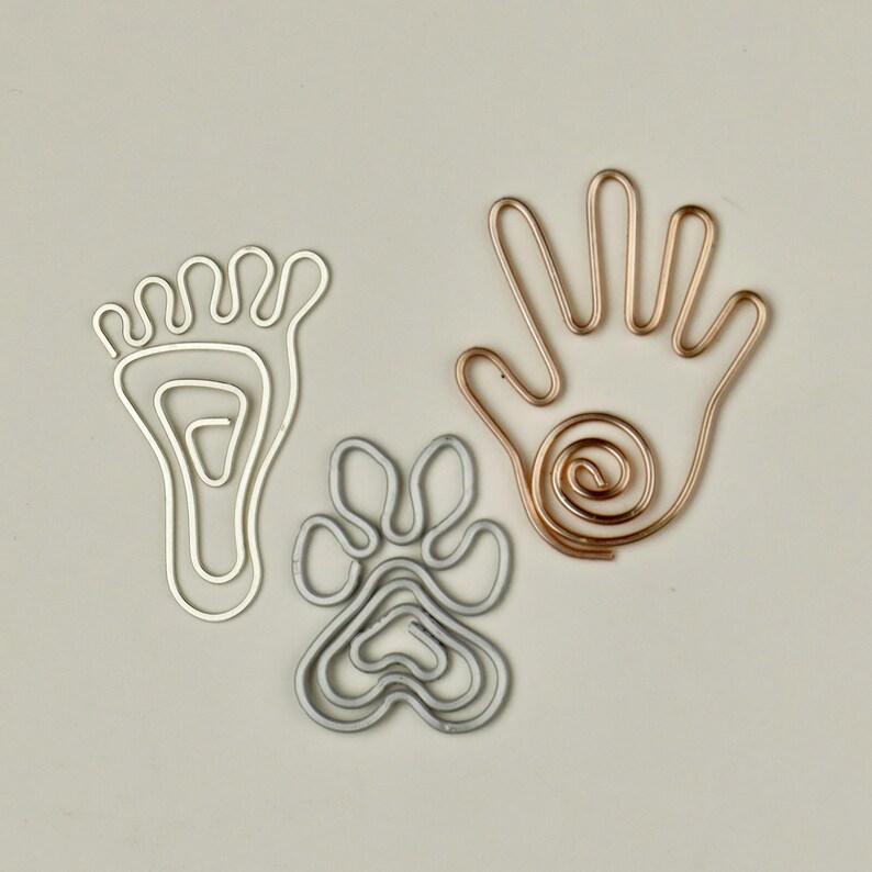 K&ouml;nnte beinhalten: Drei Drahtskulpturen, ein Fu&szlig;, ein Pfotenabdruck und eine Hand, werden auf wei&szlig;em Hintergrund gezeigt. Der Fu&szlig; und der Pfotenabdruck sind silberfarben, die Hand ist kupferfarben.