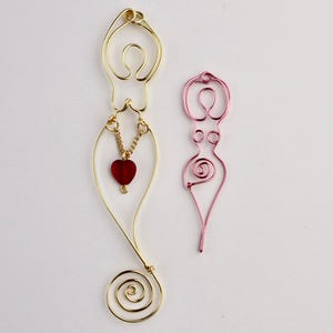May include: Two wire sculptures of a female figure. The sculpture on the left is gold and has a red heart charm hanging from it. The sculpture on the right is pink.