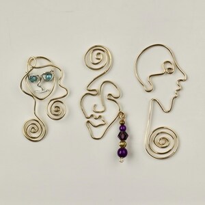 K&ouml;nnte beinhalten: Drei goldene Drahtskulpturen von Gesichtern, eine mit blauen Glasaugen und einem goldenen Drahtwirbel, eine mit einem violetten Perlenanh&auml;nger und eine mit einem einfachen Design.