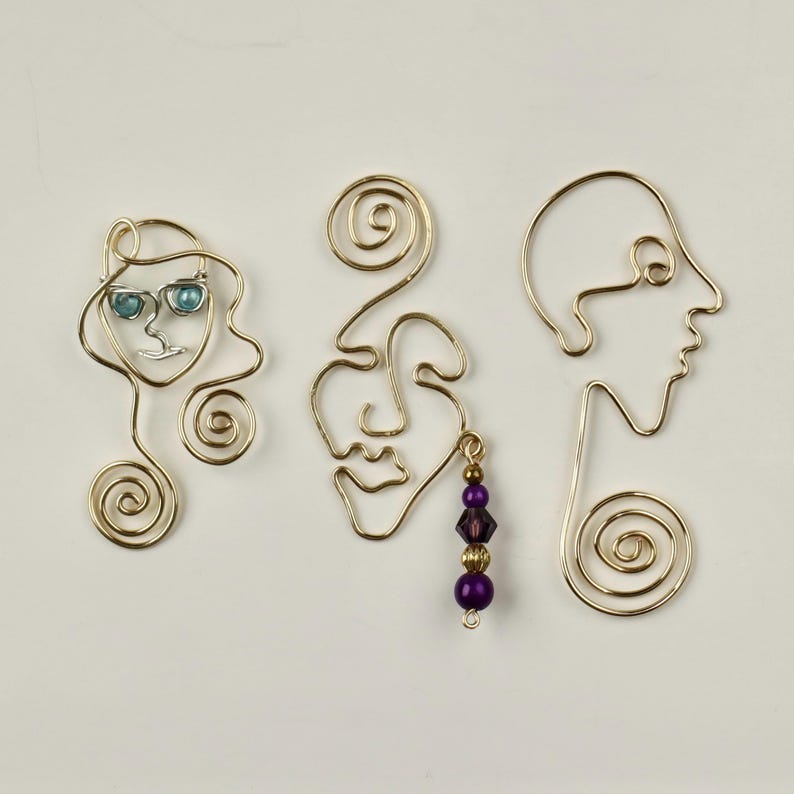 K&ouml;nnte beinhalten: Drei goldene Drahtskulpturen von Gesichtern, eine mit blauen Glasaugen und einem goldenen Drahtwirbel, eine mit einem violetten Perlenanh&auml;nger und eine mit einem einfachen Design.