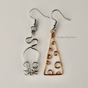 May include: A pair of silver and gold wire earrings. The left earring is shaped like a mouse and the right earring is shaped like a slice of cheese. The earrings are hanging from silver hooks.