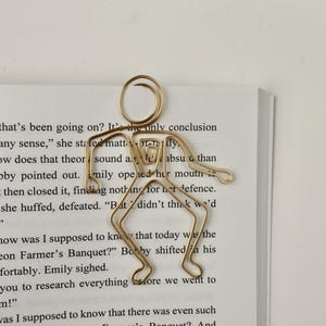 K&ouml;nnte beinhalten: Ein goldfarbenes Lesezeichen in Form einer Strichm&auml;nnchenfigur. Das Lesezeichen liegt auf einem ge&ouml;ffneten Buch mit Text &uuml;ber ein Taubenfarmer-Bankett.