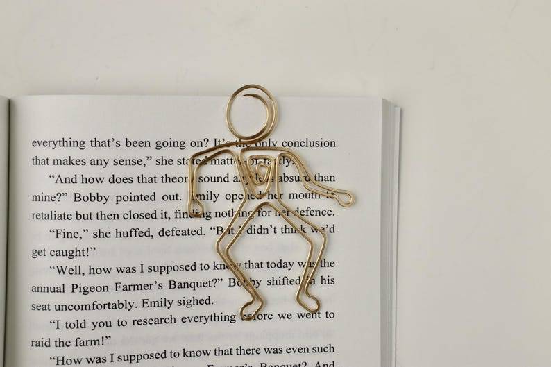 K&ouml;nnte beinhalten: Ein goldfarbenes Lesezeichen in Form einer Strichm&auml;nnchenfigur. Das Lesezeichen liegt auf einem ge&ouml;ffneten Buch mit Text &uuml;ber ein Taubenfarmer-Bankett.