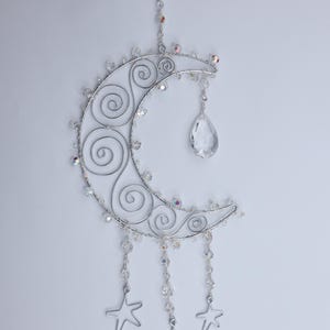 May include: A silver-toned wire crescent moon ornament featuring intricate spiral patterns, adorned with clear faceted crystal beads along the frame. A large teardrop-shaped crystal hangs from the center, with three dangling silver star charms.