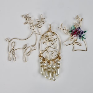 May include: Three gold-coloured wire reindeer ornaments. One is a full reindeer silhouette, another is a reindeer head with floral accents, and the third is an oval pendant with a reindeer design and dangling faux pearls.