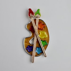 May include: A gold-toned artist's palette pin with colorful paint splotches and two crossed paintbrushes. The palette features red, orange, yellow, green, blue, and purple paint colors. The pin is a charming accessory for art lovers.