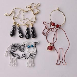 May include: Three wire sculptures of cows. One is gold with black beads, one is white with black wire and blue beads, and one is red with a red heart charm.
