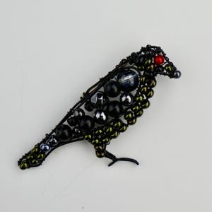 May include: A black wire bird brooch with green, black, and blue beads. The bird has a red bead for an eye.