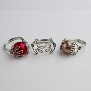 Könnte beinhalten: Drei silberne Drahtringe mit unterschiedlichen Designs. Ein Ring hat eine rote Perle, einer eine braune Perle und der andere ist ein schlichtes Drahtdesign.