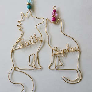 Op de afbeelding: Twee gouden draaddierornamenten, een in de vorm van een kat en de andere in de vorm van een eenhoorn. Het kattenornament heeft de naam "Sophie" erop geschreven en het eenhoornornament heeft de naam "Faith" erop geschreven. Beide ornamenten hebben kleurrijke kralen bovenaan.