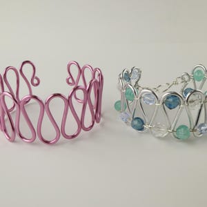 Puede incluir: Dos pulseras de alambre, una rosa y otra plateada. La pulsera plateada tiene cuentas azules y verdes.