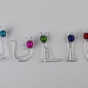 Może przedstawiać: Imię "JULIE" napisane za pomocą pięciu drucianych figurek kotów. Każdy kot ma inną kolorową szklaną koralik jako głowę: turkusowy, różowy, zielony, niebieski i czerwony.