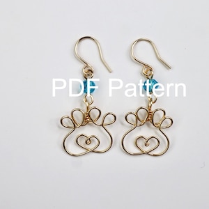 May include: A pair of gold-toned wire earrings featuring a paw print design with a heart shape at the base and a single blue bead on each hook. The text PDF Pattern is overlaid on the image. These dangle earrings have a simple wire construction.