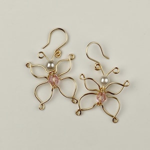 May include: A pair of gold butterfly earrings featuring pink and white beads. The earrings are crafted from wire and have a delicate design.