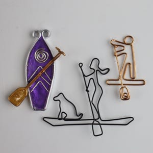 May include: A selection of wire art sculptures depicting water sports. Includes a purple and silver kayak with a gold paddle, a black wire sculpture of a person and dog on a kayak, and a gold wire sculpture of a paddleboarder.