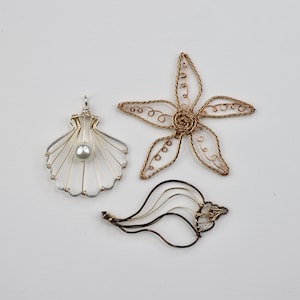 May include: Three wire art pieces: a seashell pendant with a pearl, a starfish, and a seashell. The pendant is silver and gold with a grey pearl. The starfish is gold. The seashell is black and gold.